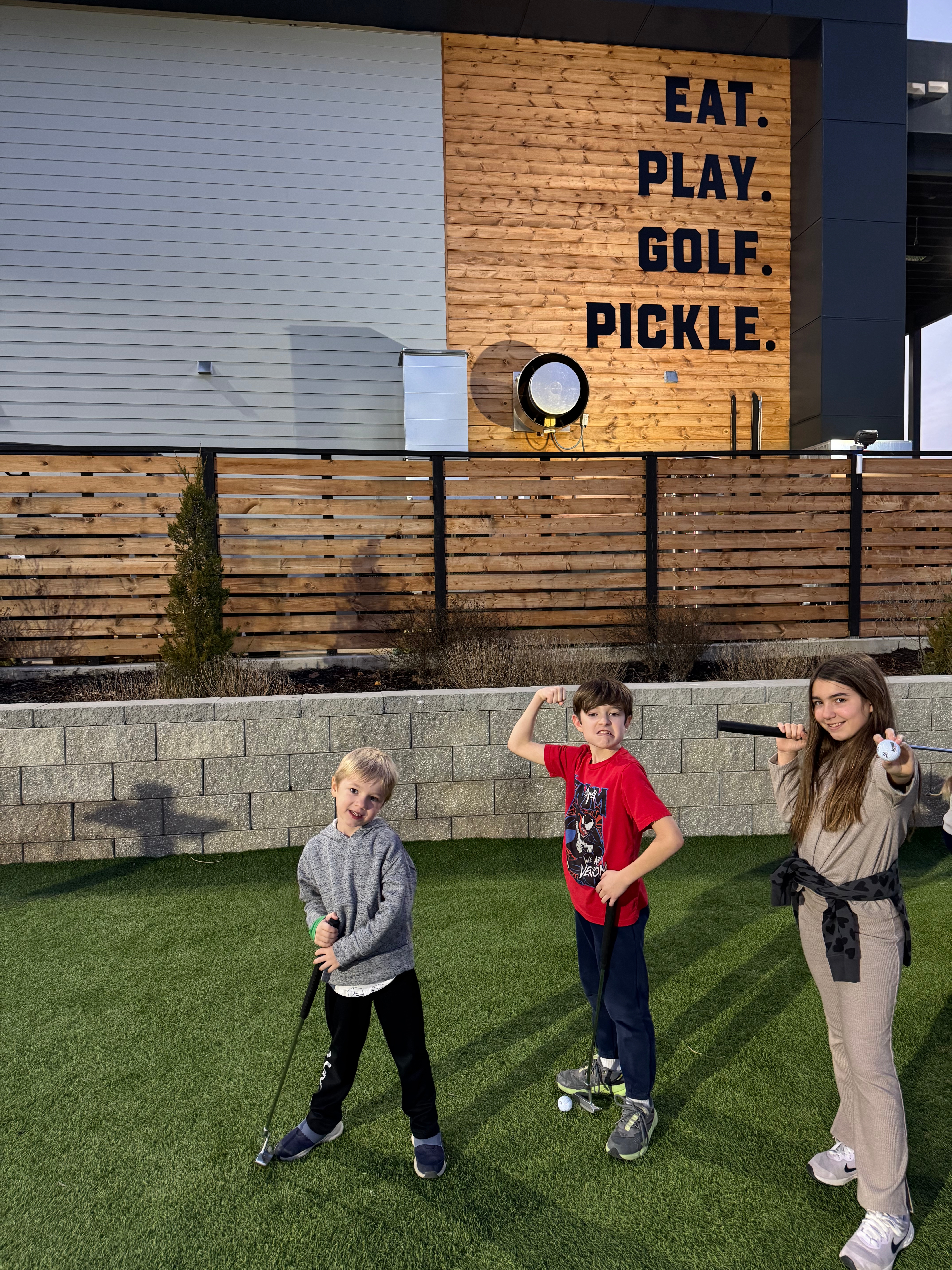 Three children pose in front of a wooden wall with the text 'EAT. PLAY. GOLF. PICKLE.' The kids stand on green grass, one holding a golf club, while the others make playful poses.