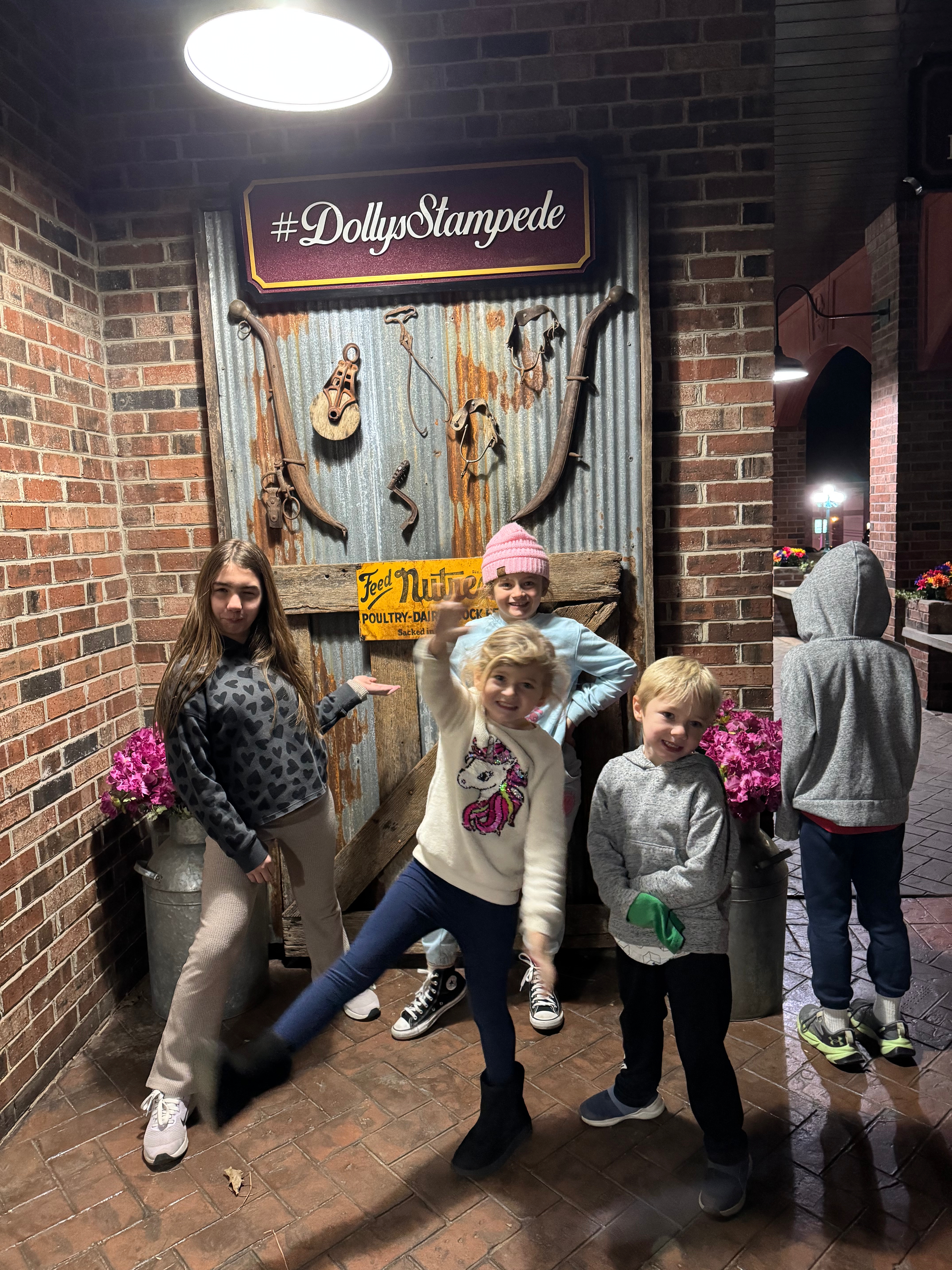 A group of children posing playfully in front of a rustic backdrop featuring a sign for Dolly Parton's Stampede dinner show, with decorative elements like Western tools and potted flowers.