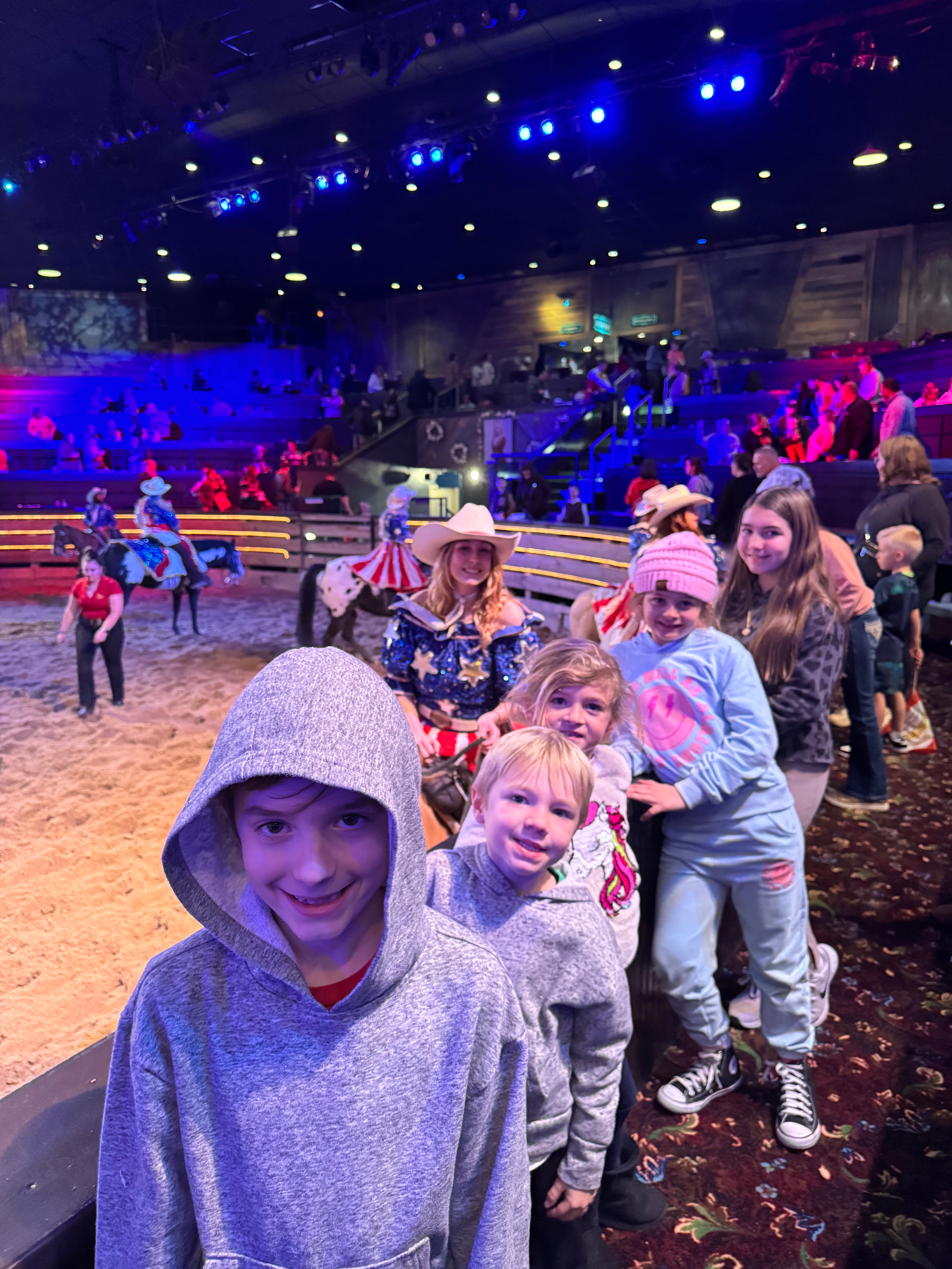 A group of children and adults, all dressed in gray hoodies, are excitedly waiting in line at a dinner show venue. Bright stage lights illuminate the background, with performers in cowboy hats and colorful costumes nearby.