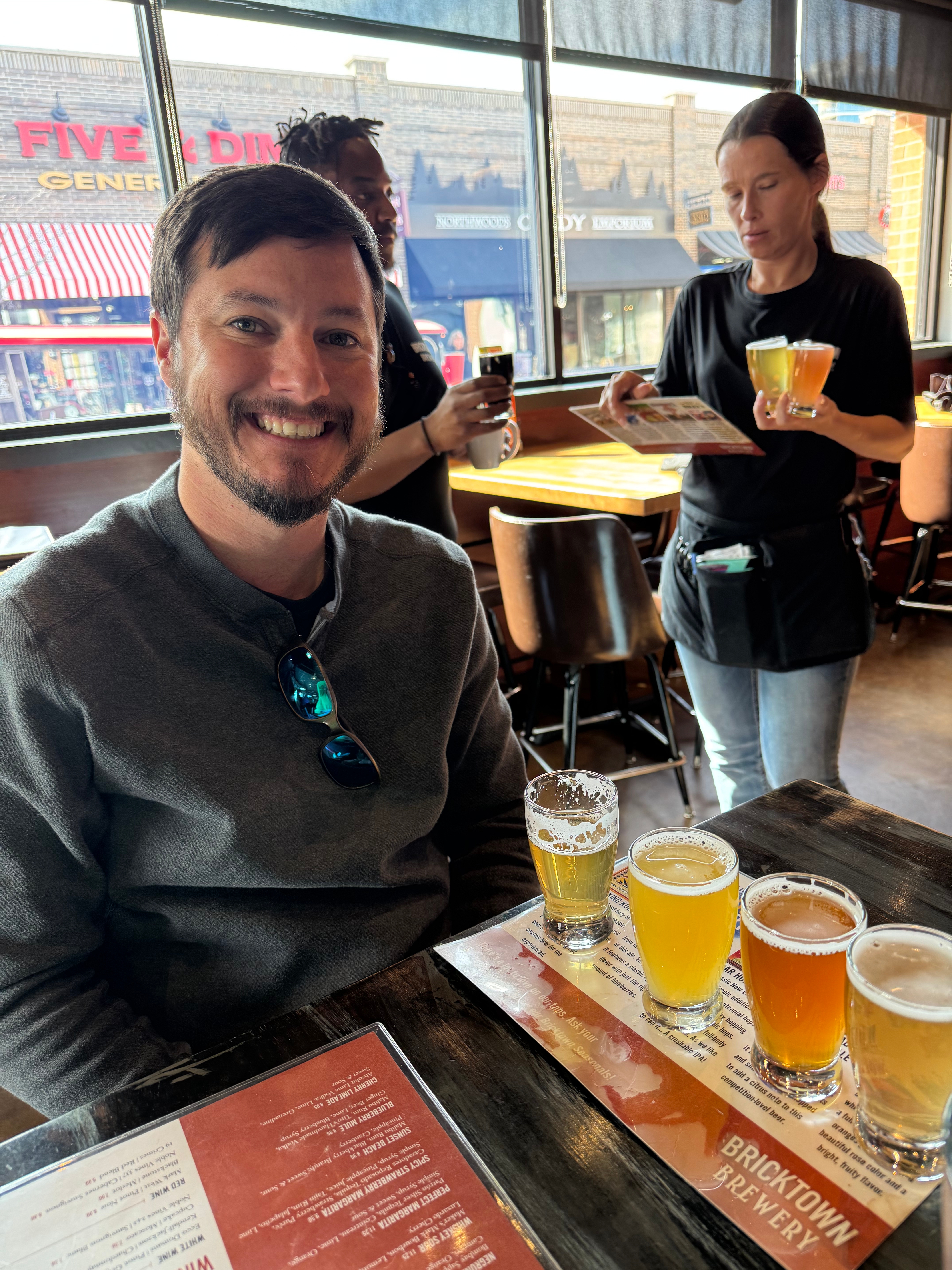 A smiling man with a beard sitting at a table in a brewery, surrounded by flight glasses of beer and a menu, with a waitress in the background holding drinks.