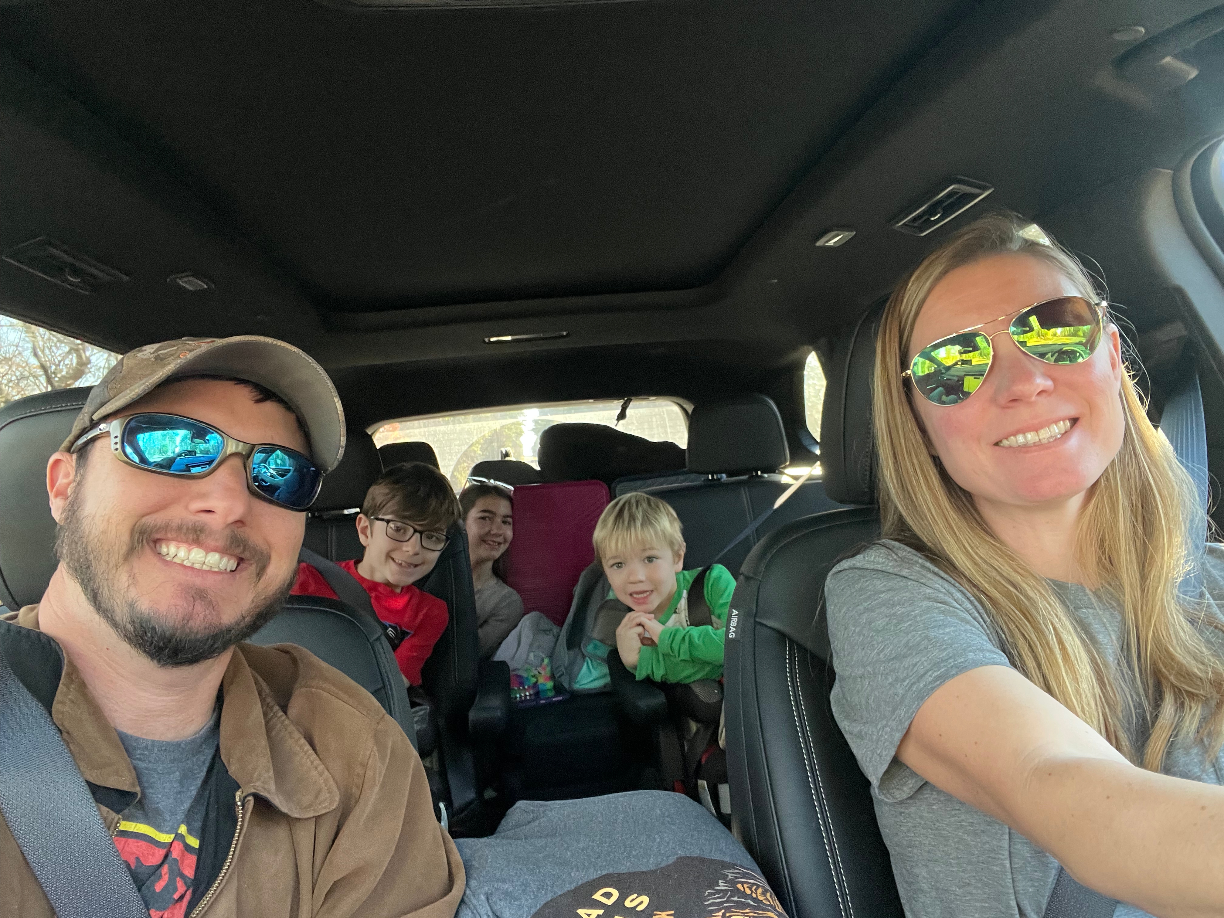 A family of five, including parents and three children, smiling for a selfie inside a car during a road trip.