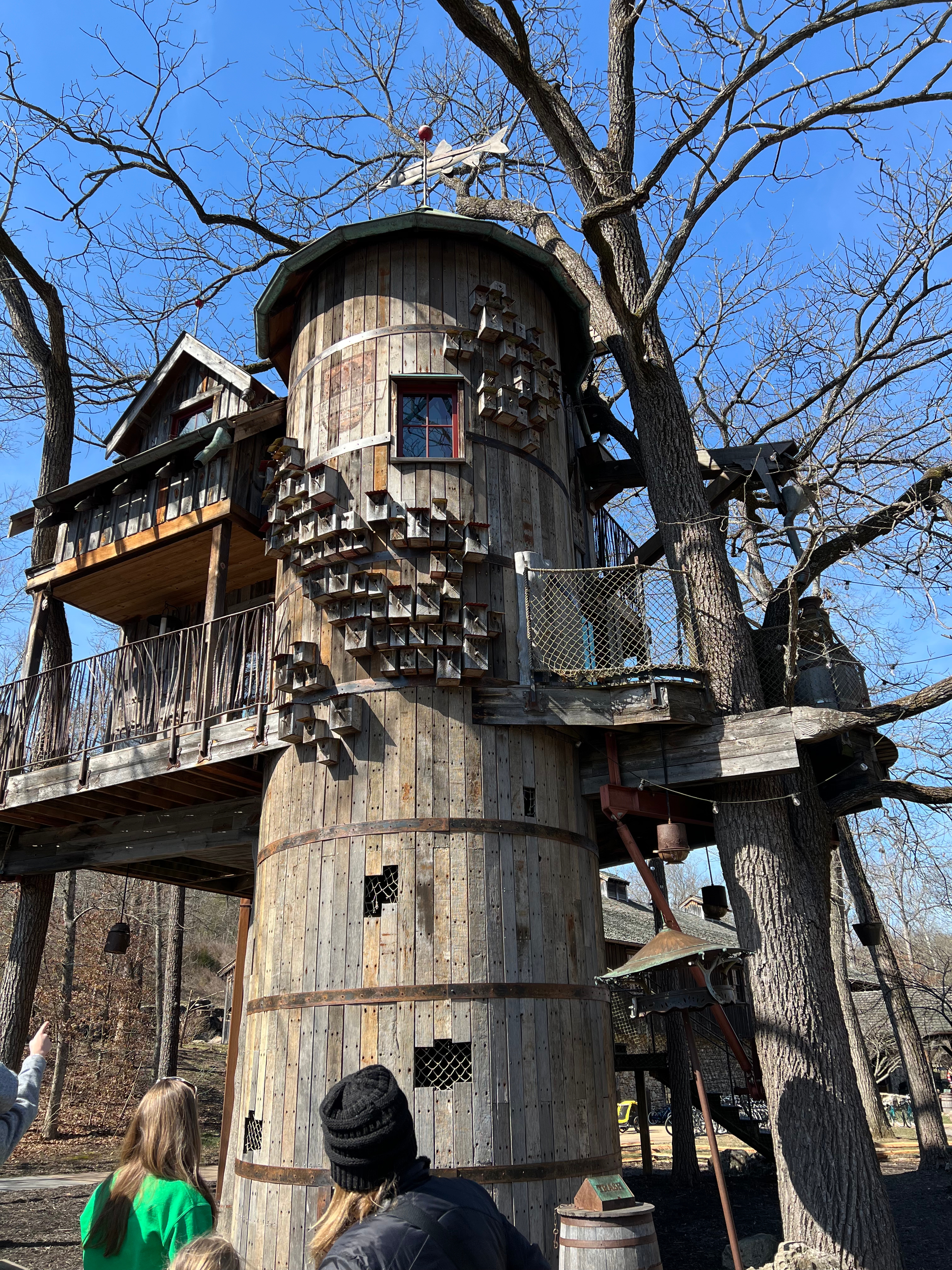 A unique treehouse structure made of wood, showcasing multiple levels, a whimsical design, and decorative elements, set against a clear blue sky with bare tree branches in the background.