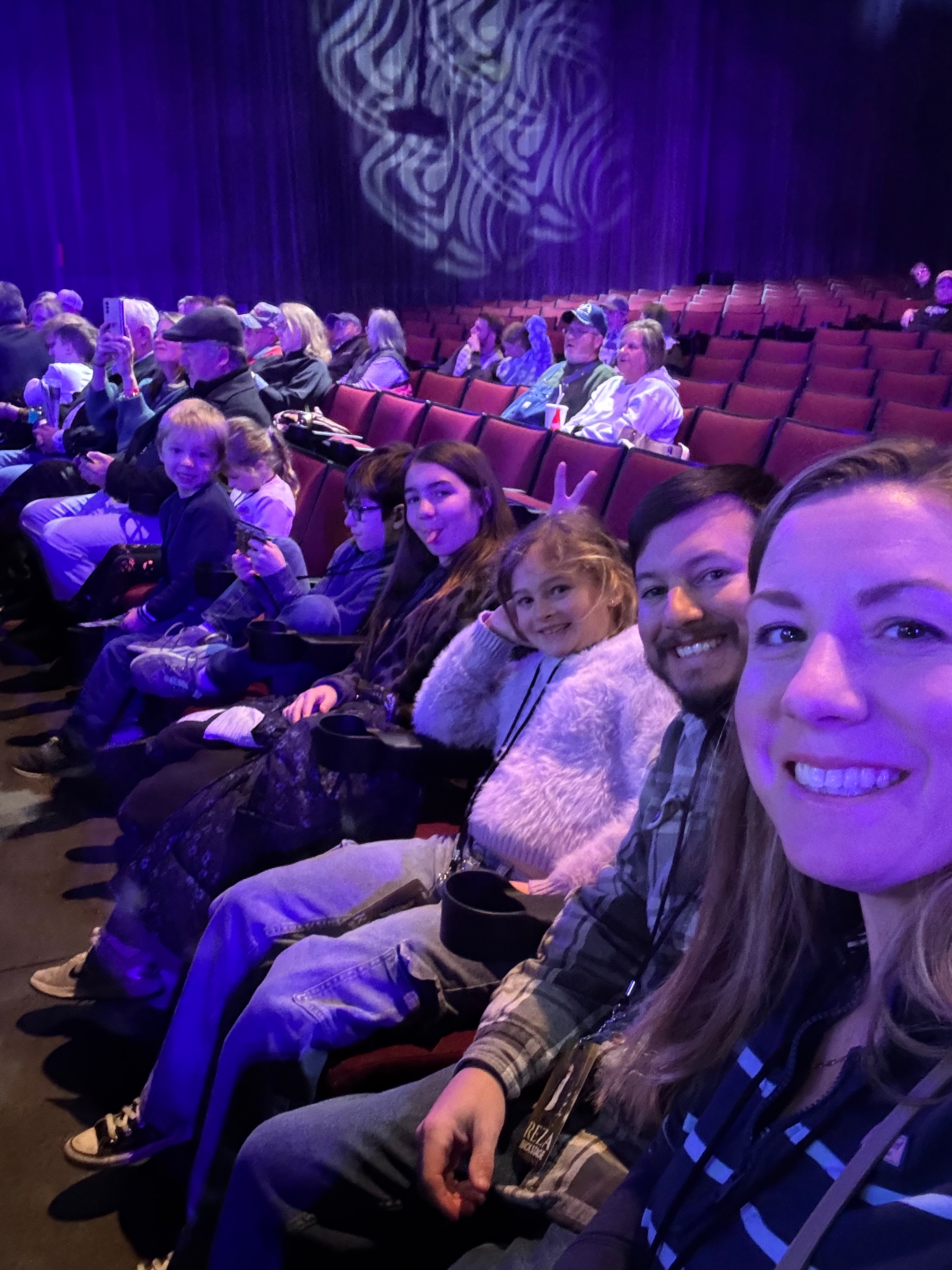 A family of five taking a selfie before a magic show, sitting in theater seats with a purple ambiance in the background.