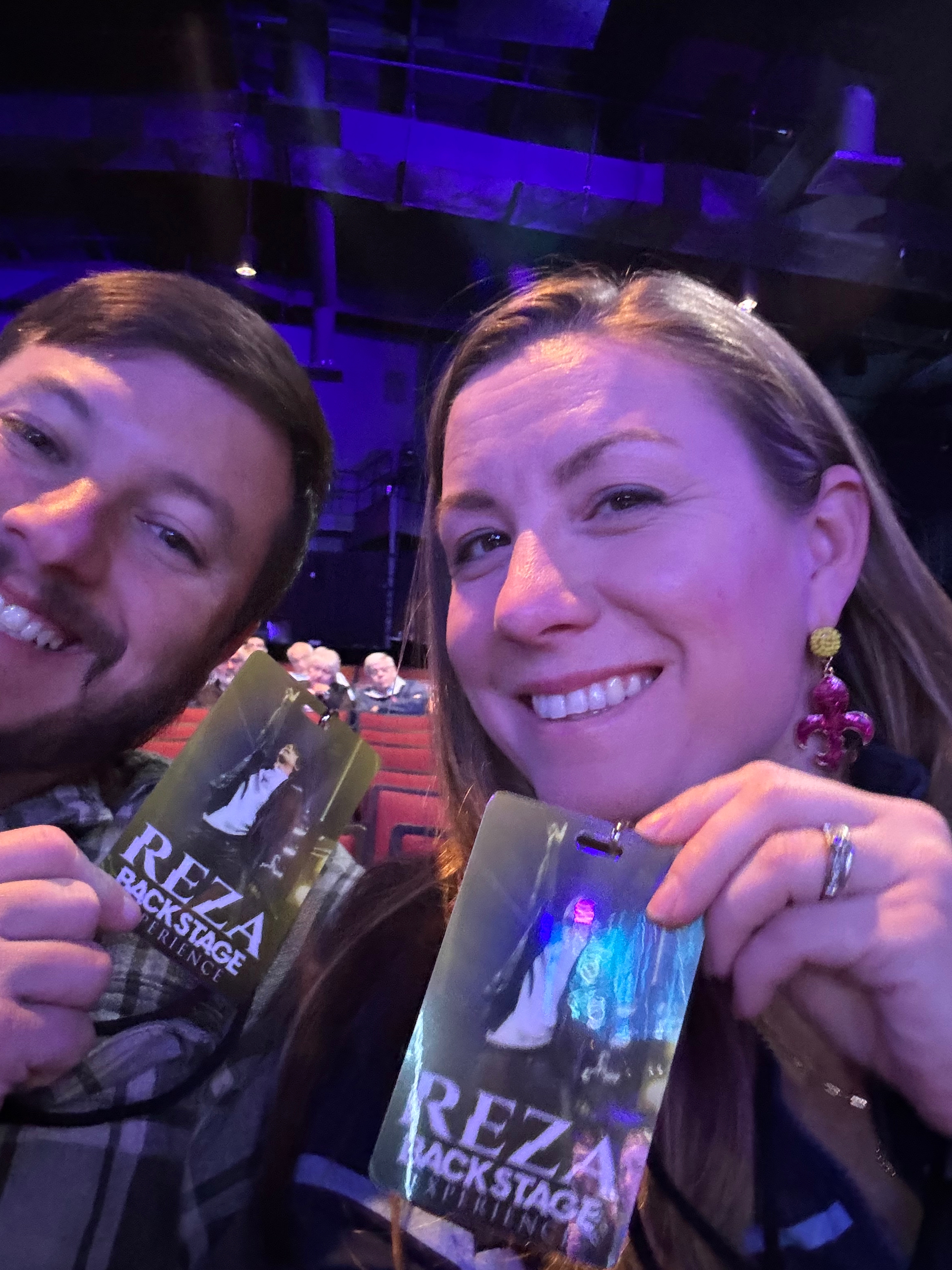 A couple smiling and holding backstage passes for Reza's magic show, seated in a theater with purple lighting in the background.