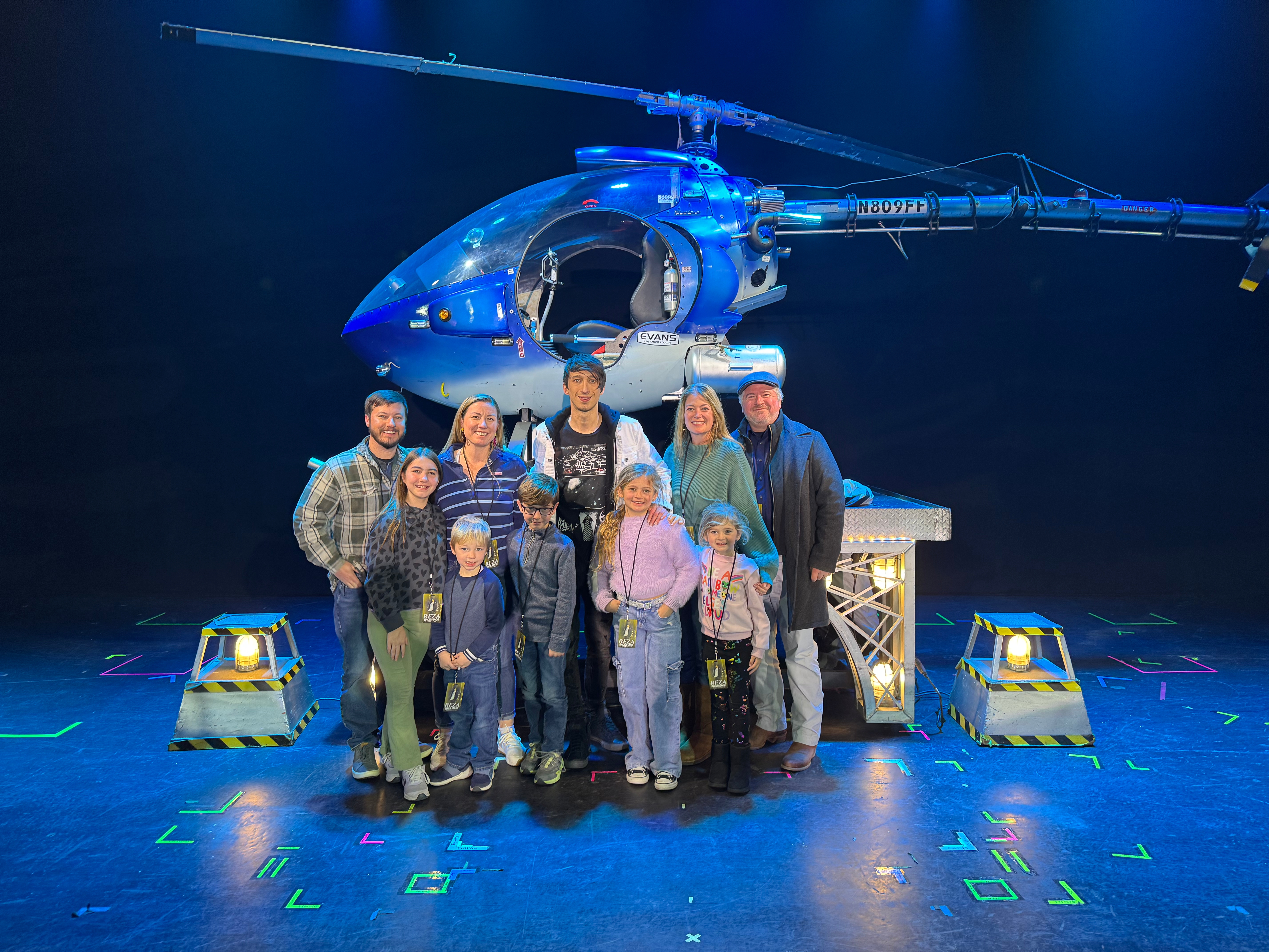 A group of adults and children posing together in front of a large helicopter prop on stage, with colorful lights in the background.