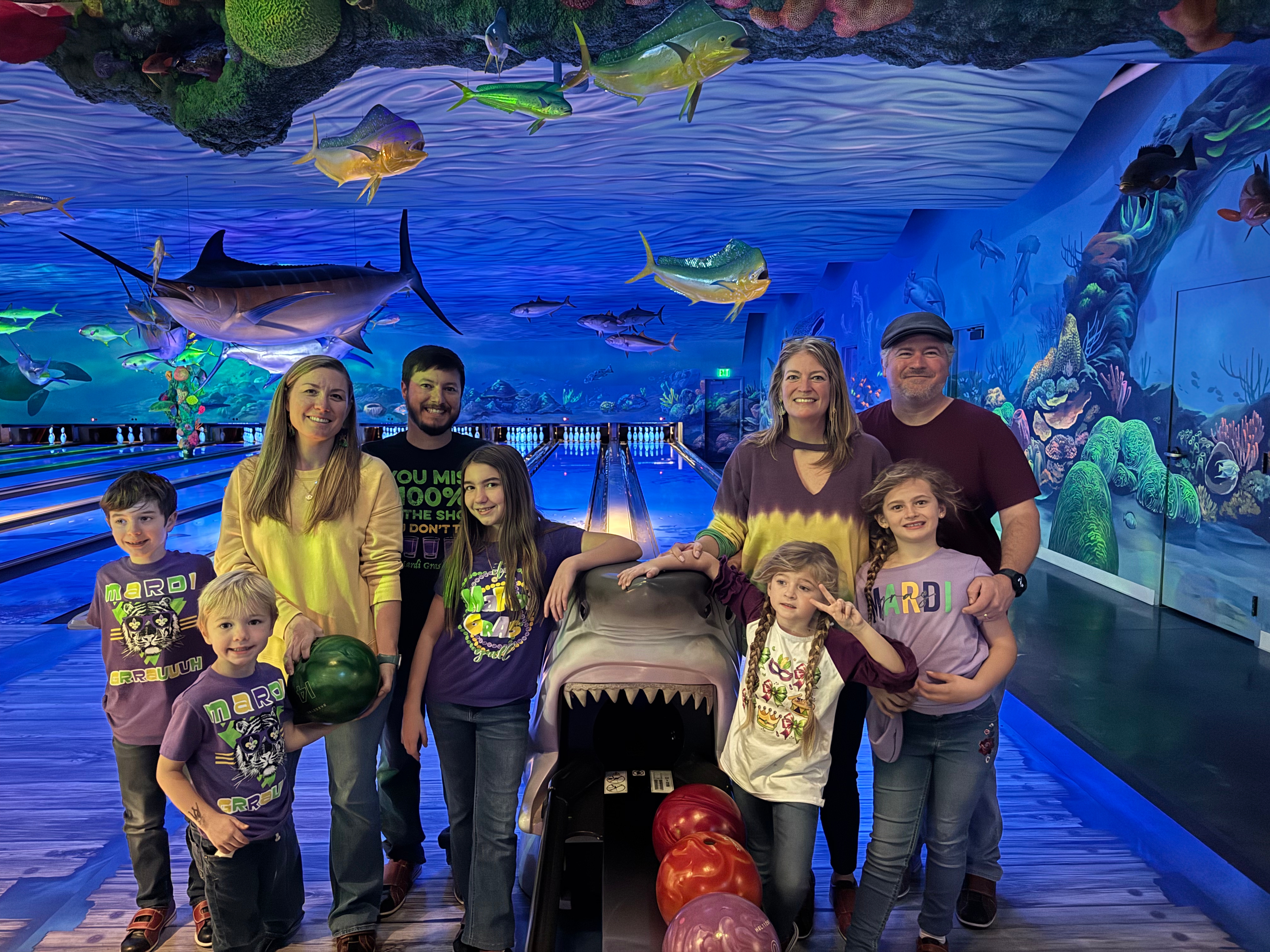 A family of five poses for a photo at a mini golf venue, surrounded by colorful underwater-themed decor, including large fish and a shark bowling ball return. The children are holding bowling balls and wearing matching 'Mardi Gras' shirts.