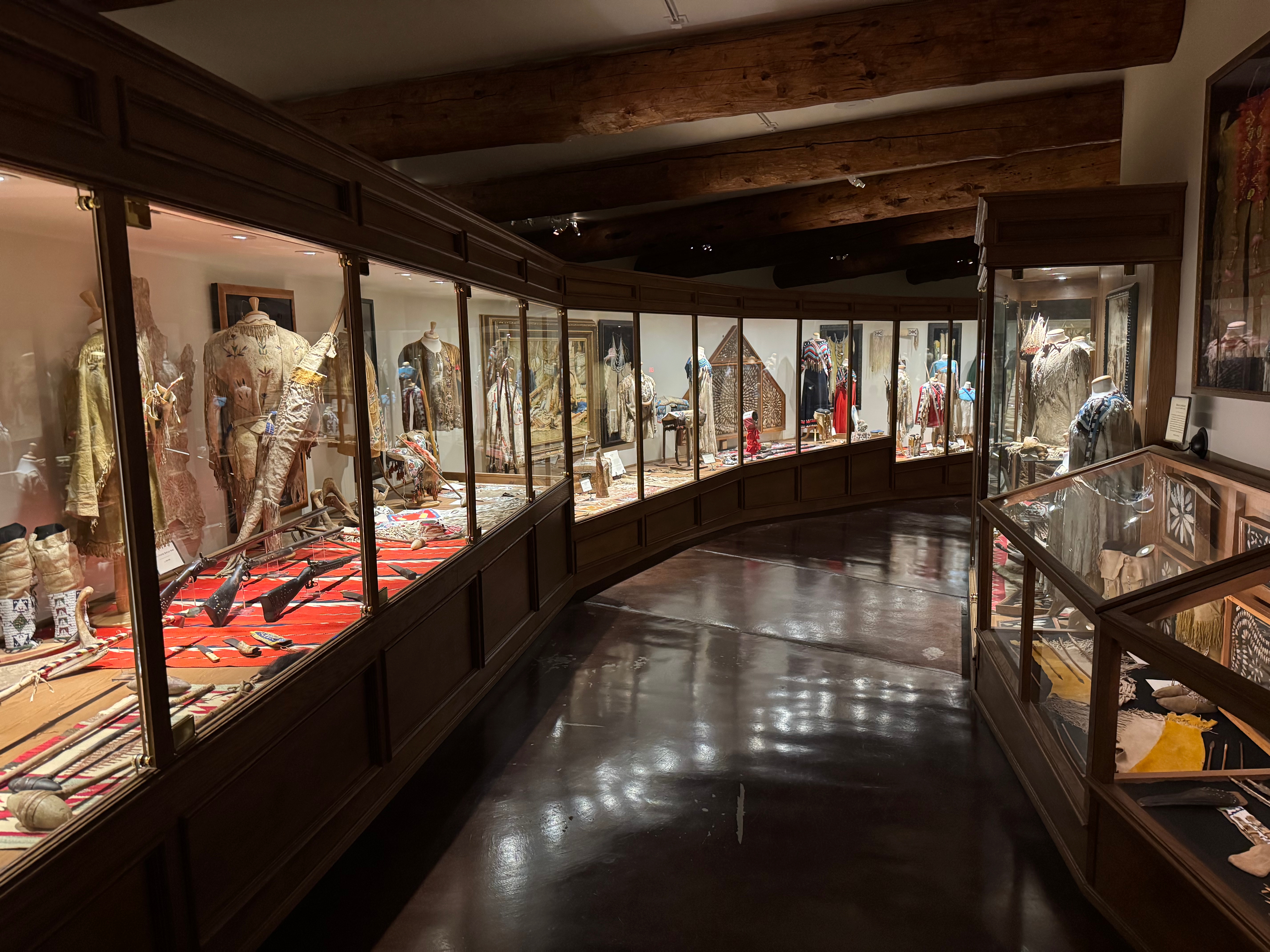 Interior view of a museum showcasing Native American artifacts, including clothing, weapons, and decorative items displayed in glass cases.