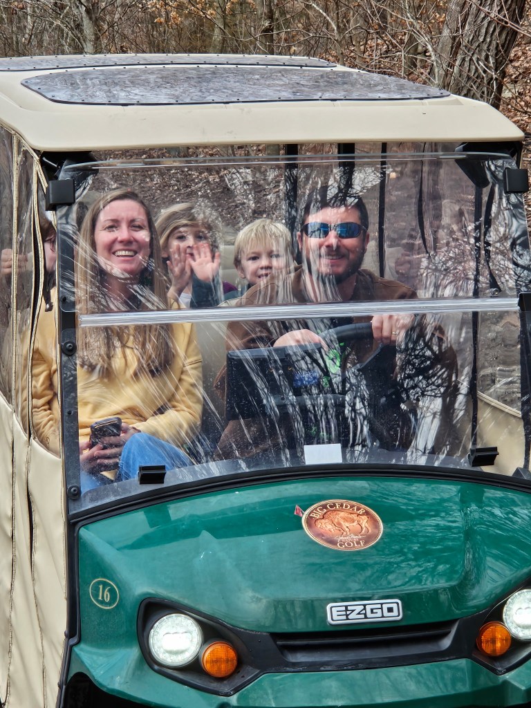 A family of five smiling in a golf cart while driving through a wooded area.