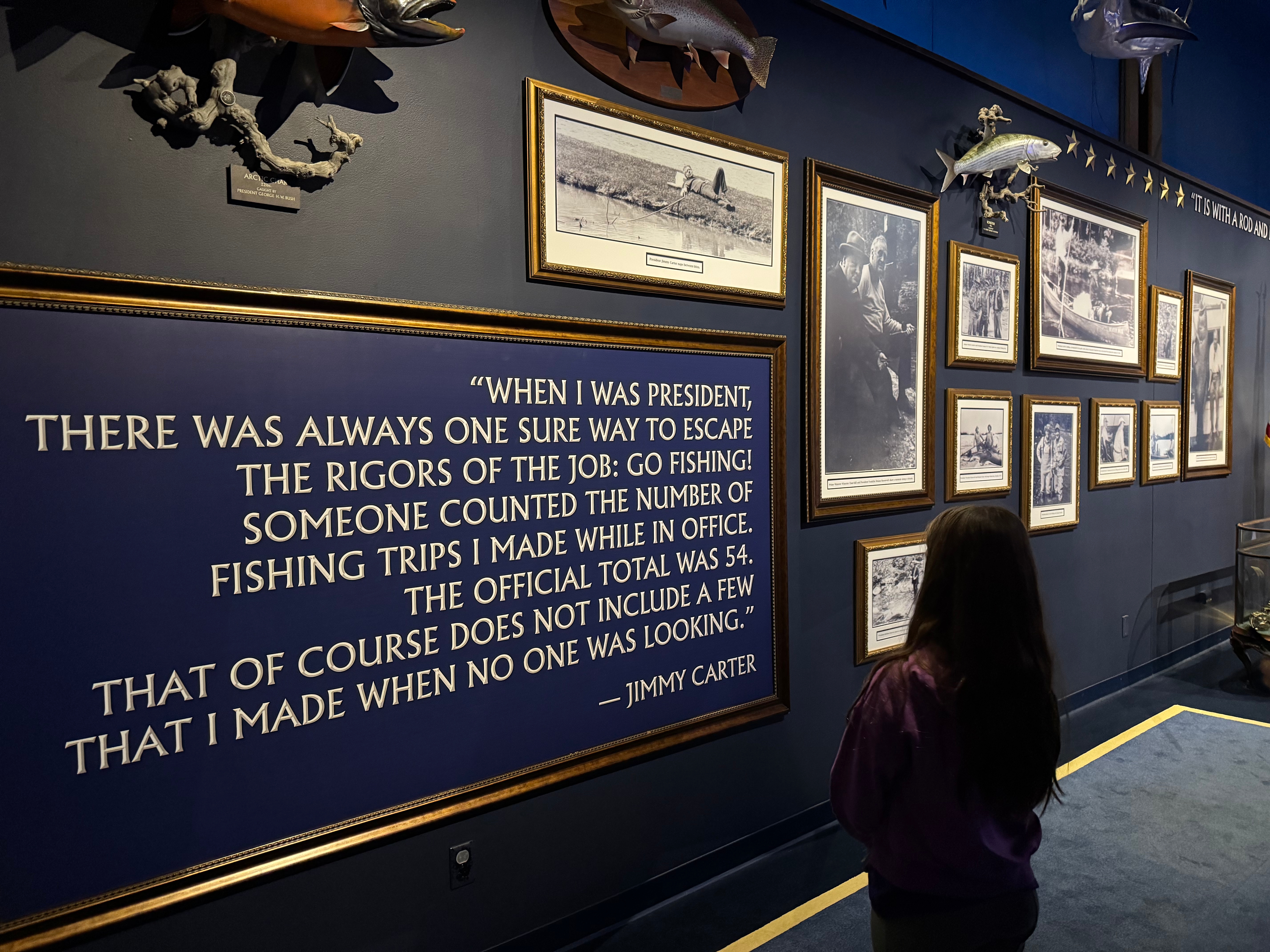 A child looking at a tribute wall with framed photographs and a quote by former President Jimmy Carter about fishing, located in a museum setting.