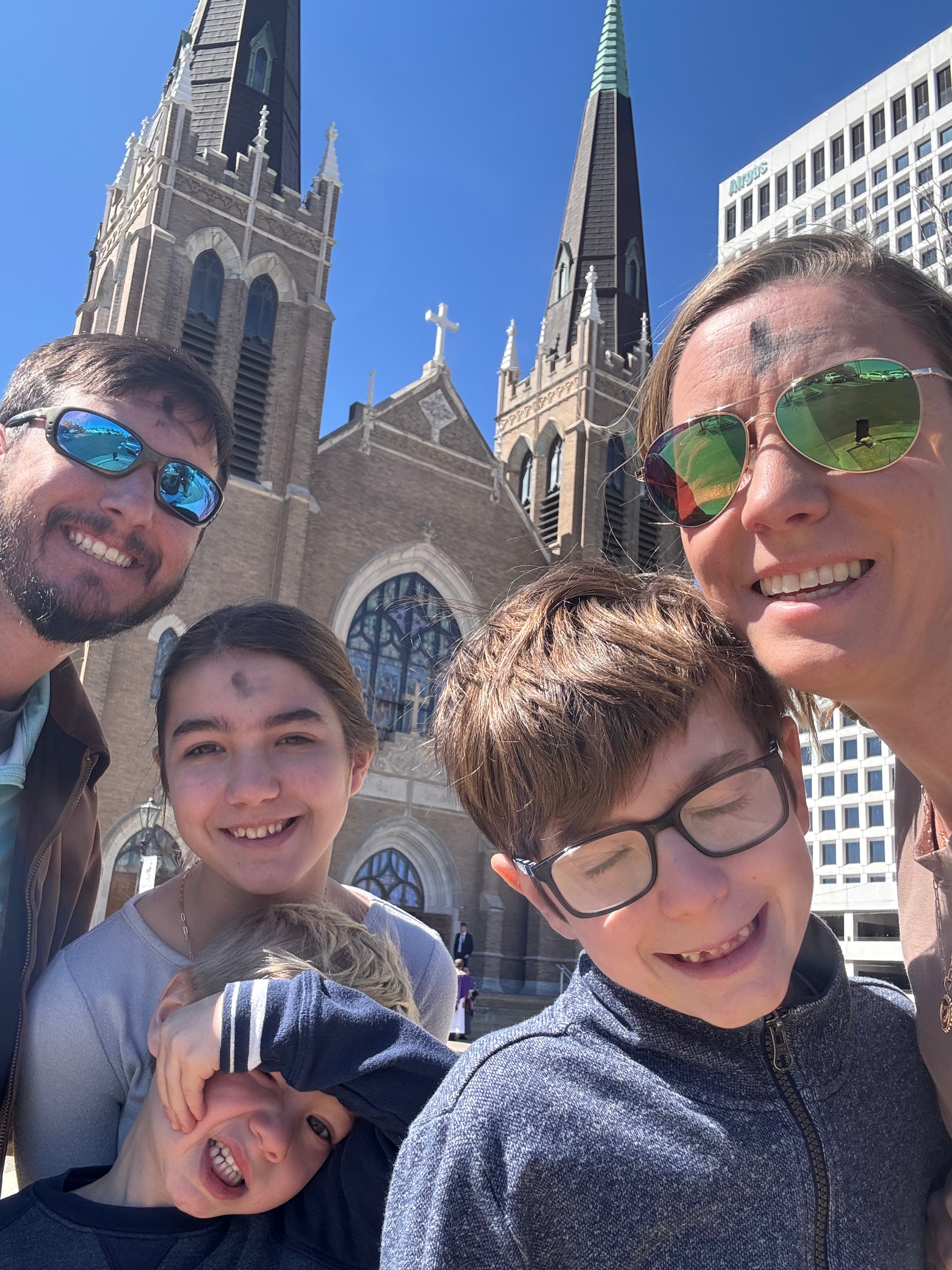 A family of five smiling together in front of a church, with visible ash crosses on their foreheads, capturing a moment after an Ash Wednesday service.