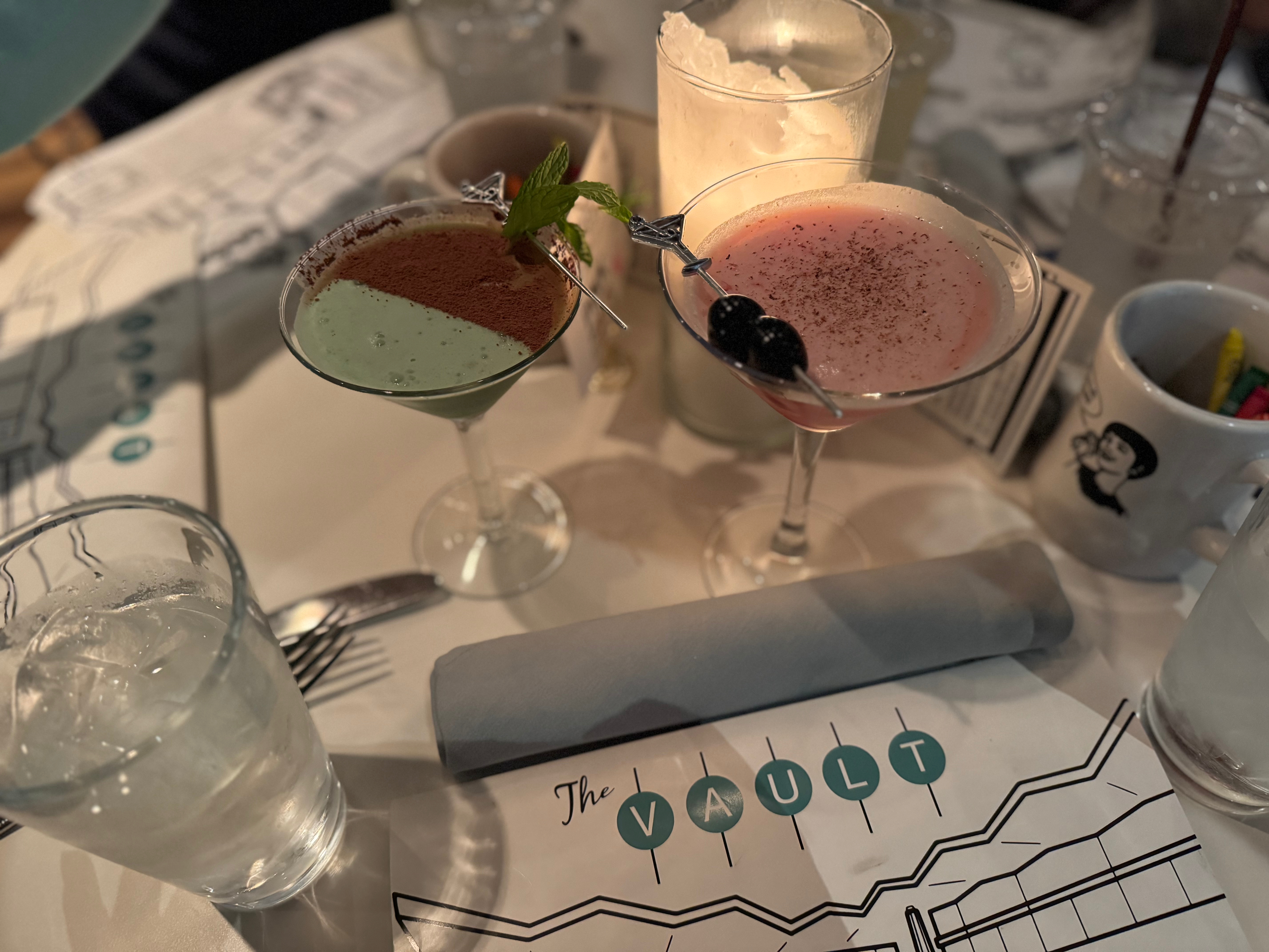 A close-up view of two cocktails on a table at The Vault restaurant, featuring a green drink garnished with mint and a pink drink with blackberries, along with a menu and glasses of water.