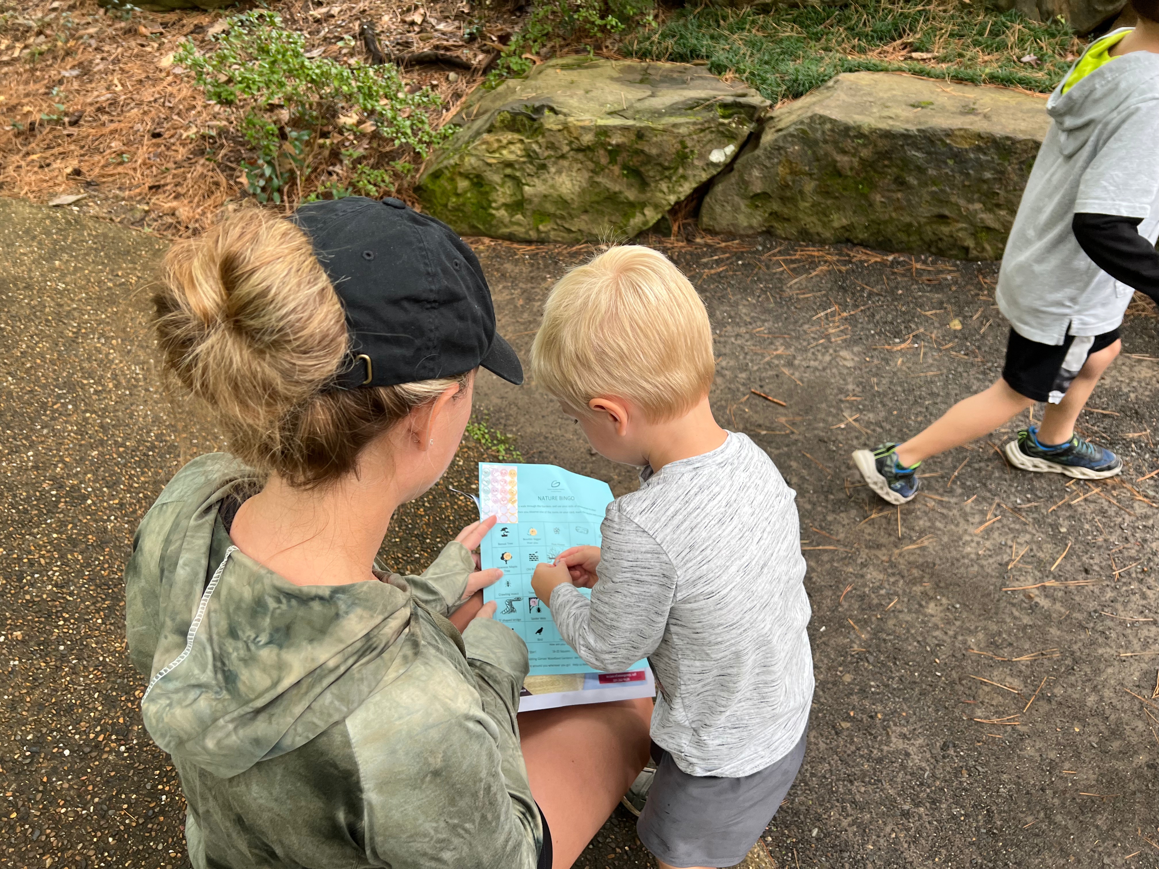 A parent and child engaging in a scavenger hunt, examining a treasure map while another child walks nearby on a path surrounded by nature.